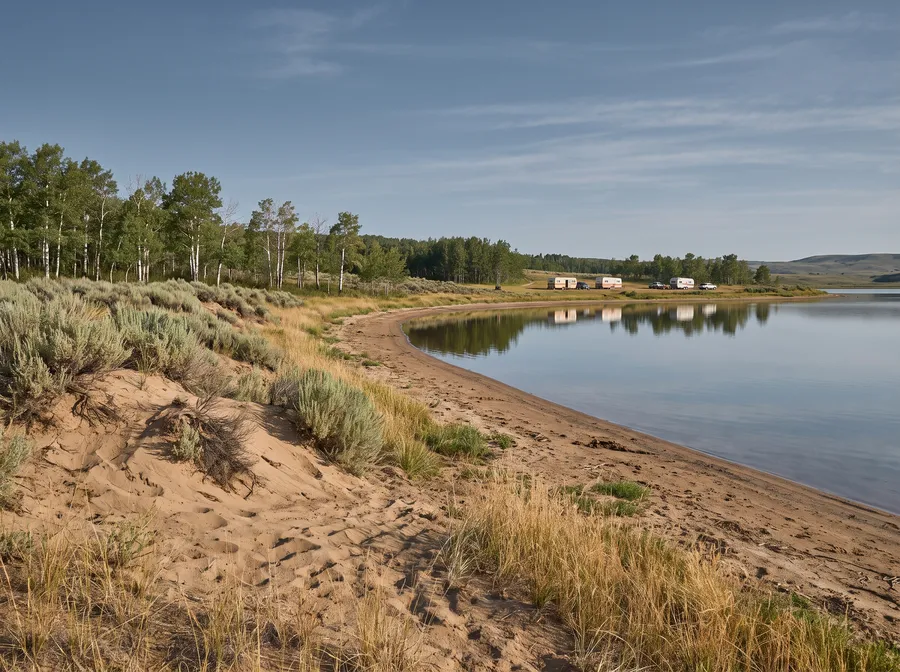 Generated scene: a calm prairie lake with a sandy shore, low dunes, aspen trees and a few travel trailers far along the shore.