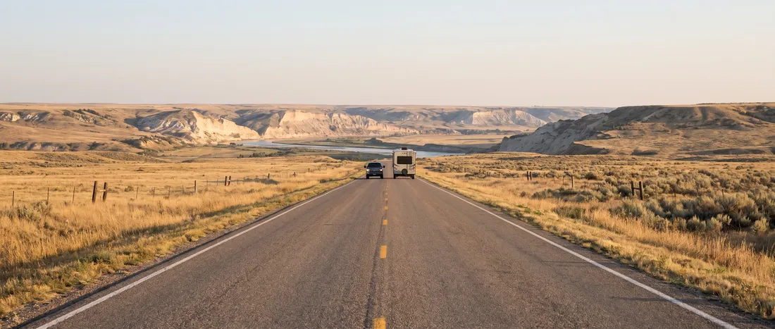 Generated scene: a two-lane prairie highway at sunrise running toward a wide river valley with pale bluffs, a pickup and a travel trailer far ahead.